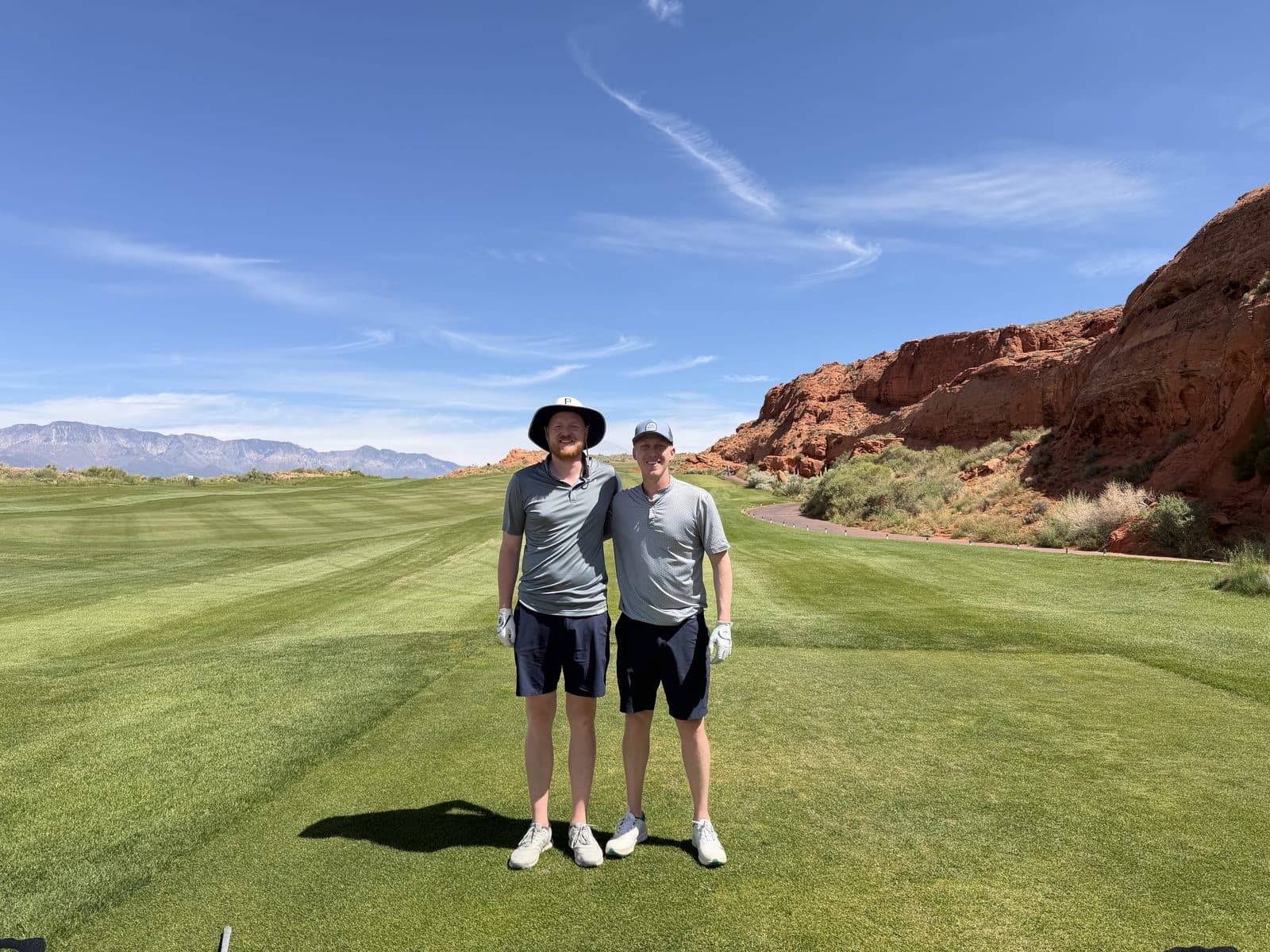 Two golfers standing on a green desert fairway near red rock cliffs.